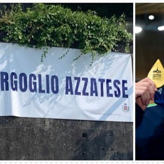 Nicolò sorridente con l'oro vivo addosso e, a sinistra, lo striscione con cui la sua Azzate lo ha celebrato questa mattina, come dopo ogni grande vittoria